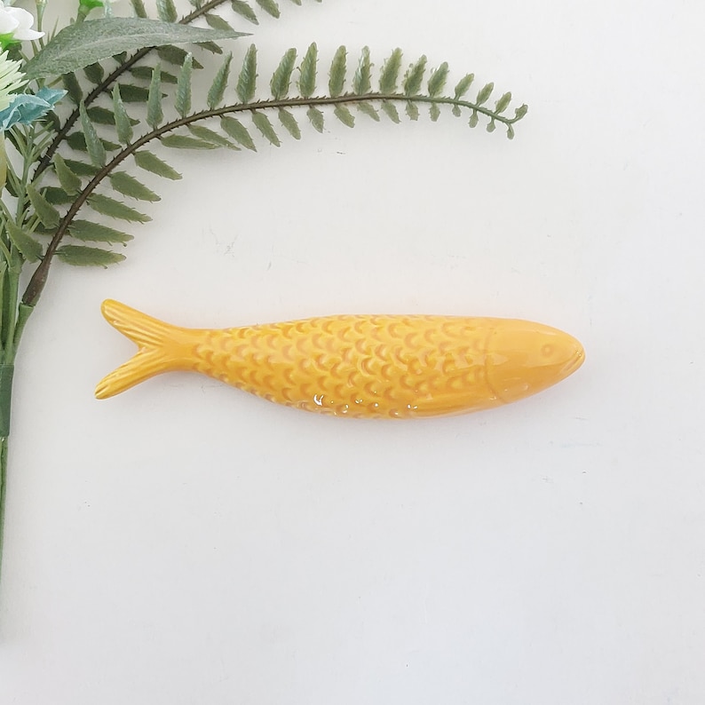 Puede incluir: Una figurita de pescado de cer&aacute;mica amarilla con una superficie texturizada. El pez tiene un cuerpo largo y delgado y una cola puntiaguda.
