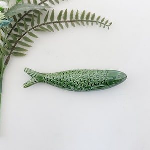Puede incluir: Una figurita de pescado de cer&aacute;mica verde con un patr&oacute;n de escamas detallado. El pez mide aproximadamente 15 cm de largo y tiene un cuerpo ligeramente curvado.