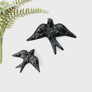 Può includere: Due rondini in ceramica nera con dettagli floreali bianchi. Le rondini sono in volo, con le ali spiegate. La rondine più grande è lunga circa 20 cm, e la più piccola è lunga circa 13 cm. Su sfondo bianco.