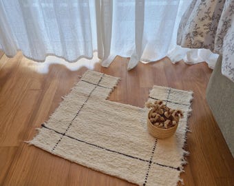 May include: A cream-colored, textured rug with a unique shape and dark blue grid pattern. The rug has a fringed edge and sits on a wooden floor. A small woven basket with dried flowers is on the rug.
