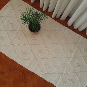 May include: A white rug with a diamond pattern and a raised texture. The rug is made of a soft, fluffy material and has a subtle sheen. A small potted plant is sitting on the rug.