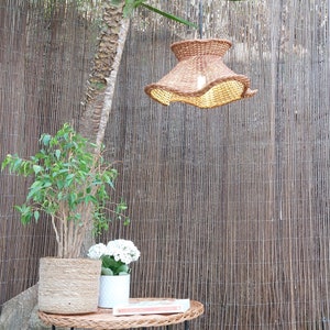 May include: A woven rattan pendant light with a ruffled edge hangs above a small wicker table. A potted plant and a white flower pot sit on the table. A tall tree trunk and a bamboo fence are in the background.