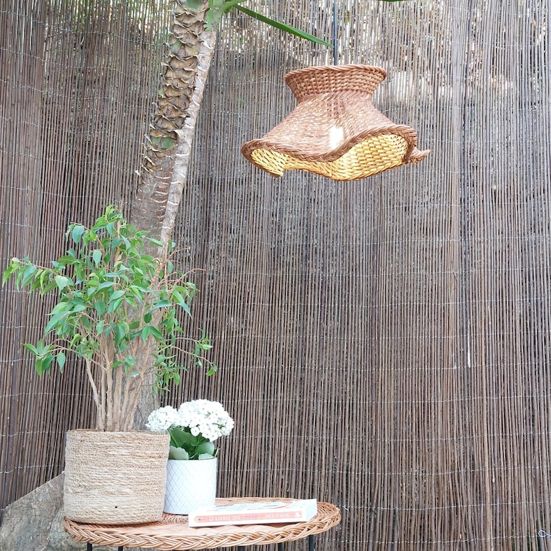 May include: A woven rattan pendant light with a ruffled edge hangs above a small wicker table. A potted plant and a white flower pot sit on the table. A tall tree trunk and a bamboo fence are in the background.