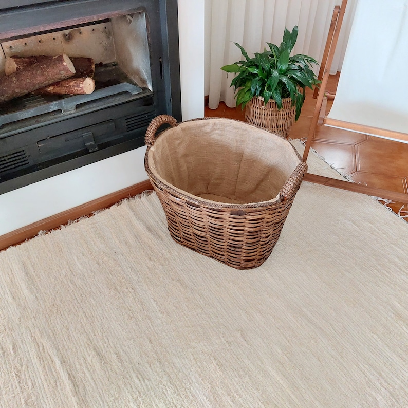 May include: A woven wicker basket with a burlap lining and two handles. The basket is brown and sits on a cream-colored rug. A fireplace with logs is in the background.