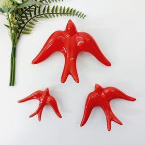 Könnte beinhalten: Drei glänzend rote Keramikschwalben in verschiedenen Größen werden vor einem weißen Hintergrund gezeigt. Die Vögel haben ausgebreitete Flügel und sind so angeordnet, dass sie einen Flug suggerieren. Eine grüne Pflanze mit kleinen Blättern befindet sich in der oberen linken Ecke.