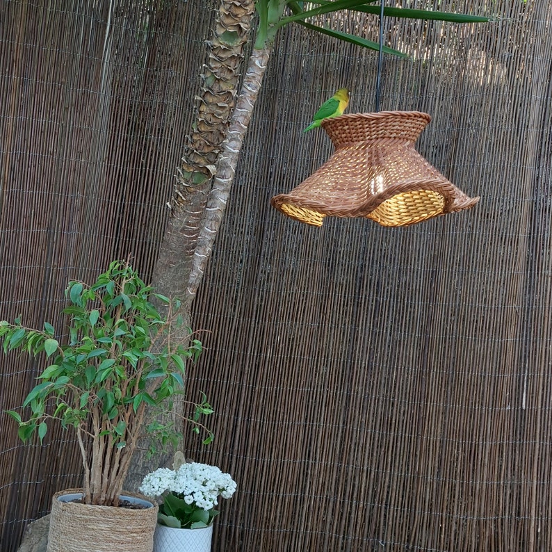 May include: A woven brown pendant light with a flared shade hangs from a black cord. A small green and yellow bird perches on top of the light fixture. The background includes a bamboo fence, a tree trunk, and potted plants.