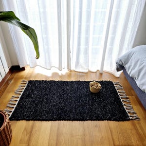 May include: A rectangular black rug with a textured weave and fringed edges. The rug is placed on a hardwood floor, with a small woven basket containing dried flowers. A large green plant and white curtains are in the background.