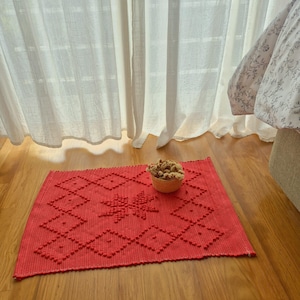 May include: A rectangular red woven mat featuring a raised diamond pattern and a central star-like motif. A small, round, light brown woven basket filled with dried natural-coloured botanical elements sits on the surface of the red mat.