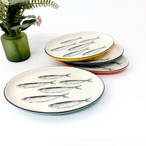 Puede incluir: Una pila de cuatro platos ovalados de cerámica con una superficie de color crema que presenta un patrón de sardinas en blanco y negro. Cada plato tiene un borde de color distinto, incluyendo amarillo, azul, naranja y azul claro, con un contorno negro.