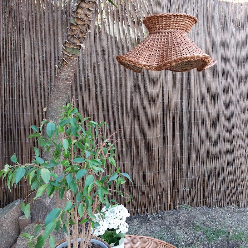May include: A woven brown lampshade with a flared, ruffled design hangs from above. The lampshade is made of natural wicker material. The background features a bamboo fence, a tree trunk, and green plants in a pot.