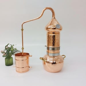Puede incluir: Un alambique de cobre con acabado martillado, con un tubo condensador curvado y un termómetro. El alambique tiene una columna cilíndrica y una base redonda con asas. Una pequeña olla de cobre está conectada.