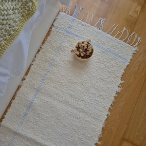 Peut inclure: Tapis rectangulaire blanc cassé avec des rayures bleues et des bords effilochés. Un petit panier en osier rempli de fleurs séchées est posé sur le tapis. Le tapis est en matière texturée, posé sur un plancher en bois.