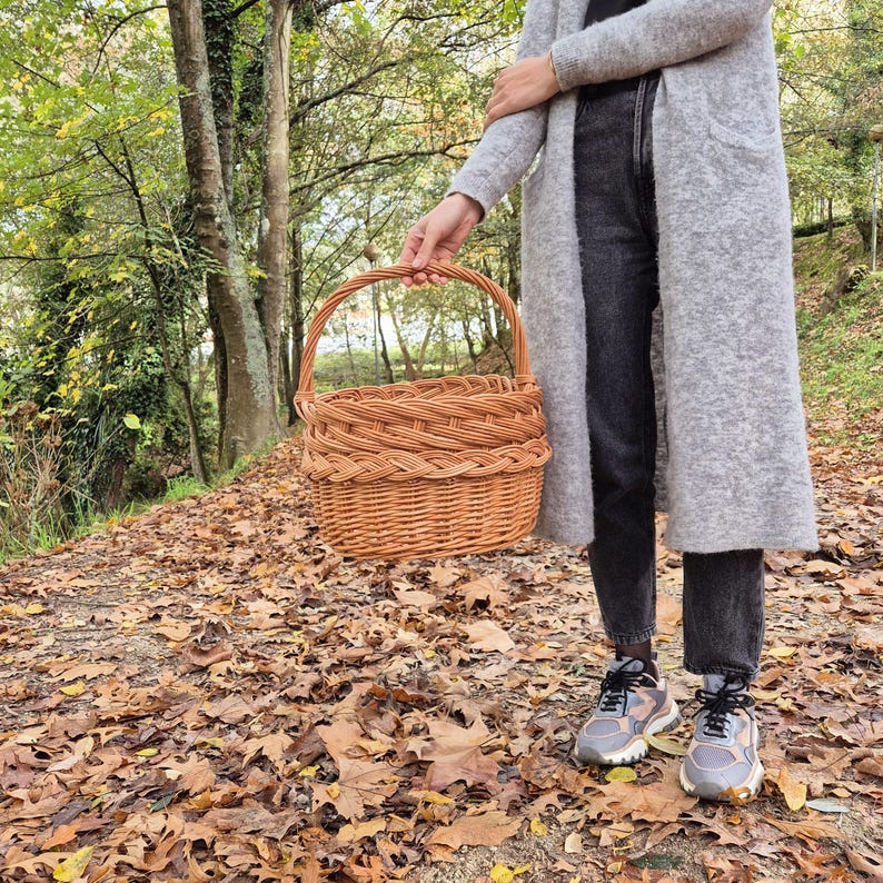May include: A woven wicker basket with a handle, held by a person in a gray cardigan and jeans, standing in a forest setting with fallen leaves on the ground.