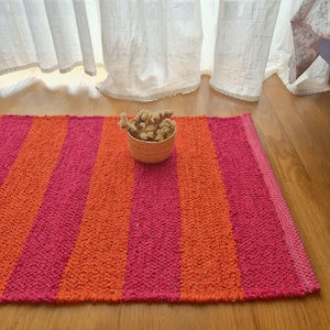 Puede incluir: Una alfombra rectangular con rayas verticales rosas y naranjas. Una pequeña cesta tejida llena de flores secas está colocada encima. La alfombra está sobre un suelo de madera, con cortinas blancas al fondo.