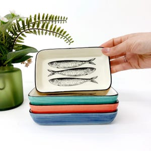Pode incluir: Um conjunto de pratos retangulares de cer&acirc;mica em tons de azul, coral e verde-azulado. O prato superior &eacute; branco sujo com uma borda preta e apresenta tr&ecirc;s ilustra&ccedil;&otilde;es de peixes em preto e branco. Decora&ccedil;&atilde;o de casa.