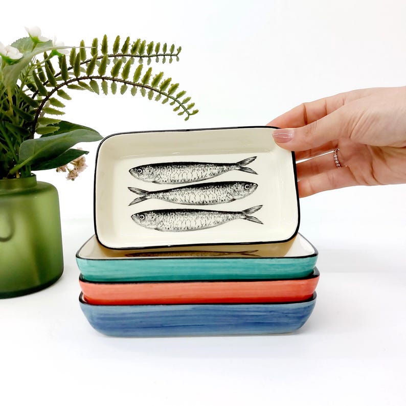 Pode incluir: Um conjunto de pratos retangulares de cer&acirc;mica em tons de azul, coral e verde-azulado. O prato superior &eacute; branco sujo com uma borda preta e apresenta tr&ecirc;s ilustra&ccedil;&otilde;es de peixes em preto e branco. Decora&ccedil;&atilde;o de casa.