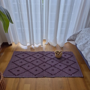 Può includere: Un tappeto rettangolare viola con un motivo a rombi e un bordo bianco è appoggiato su un pavimento in legno. Un piccolo cestino intrecciato pieno di fiori secchi è sul tappeto. Tende bianche sullo sfondo.
