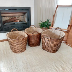 May include: Three woven wicker baskets in varying sizes, each with a burlap lining and sturdy handles. The baskets are a warm brown color, perfect for storage or decorative use. They are arranged on a cream-colored rug.