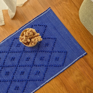 May include: A rectangular blue rug featuring a raised diamond pattern created with small pom-poms. A small, round, light-colored woven basket filled with dried, textured botanical elements sits on the surface of the rug.