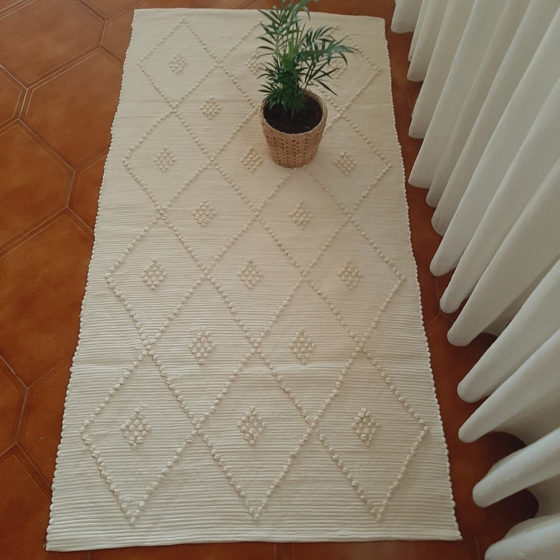 May include: A white woven rug with a diamond pattern and raised pom-poms. The rug is on a brown tiled floor.