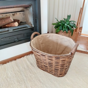 May include: A large, oval-shaped wicker basket with a burlap liner and woven handles. The basket is brown and sits on a cream-colored rug, near a fireplace. The basket is designed for storing firewood.