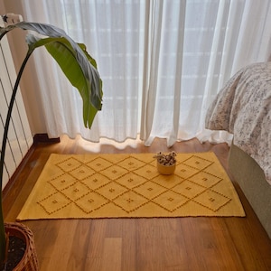 Puede incluir: Alfombra rectangular de color amarillo mostaza con un diseño de rombos y un borde festoneado. La alfombra está colocada sobre un suelo de madera cerca de una ventana con cortinas blancas. Un pequeño cuenco con flores secas está sobre la alfombra.