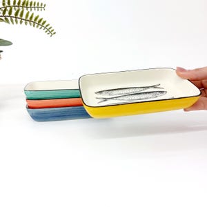 Pode incluir: Uma pilha de pratos retangulares de cer&acirc;mica em v&aacute;rias cores. O prato superior &eacute; amarelo com uma borda preta e apresenta uma ilustra&ccedil;&atilde;o em preto e branco de dois peixes. Outros pratos s&atilde;o verde-azulado, laranja e azul.