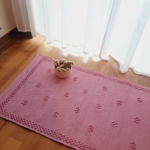 Pu&ograve; includere: Un tappeto rettangolare rosa con una superficie strutturata e un motivo a pois. Il tappeto ha un bordo di piccoli punti in rilievo. Un piccolo cestino intrecciato pieno di fiori secchi &egrave; appoggiato sul tappeto.