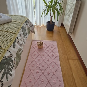 Puede incluir: Una alfombra rectangular de color rosa claro con un diseño de rombos y bolitas texturizadas. La alfombra está colocada sobre un suelo de madera cerca de una cama con una colcha estampada en verde y blanco. Una pequeña cesta de flores secas está sobre la alfombra.