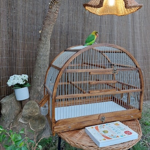 May include: A wooden birdcage with a white interior and a metal wire mesh. The cage has a rounded top and a small door on the side. A green and yellow parakeet is perched on top of the cage.