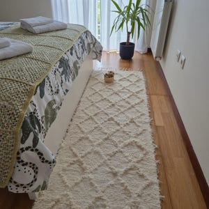 Peut inclure: Un tapis rectangulaire crème avec un motif en losanges est posé sur un parquet. Le tapis est placé à côté d'un lit avec une couverture à motifs et une couverture en crochet. Une petite plante et un bol en bois sont également visibles.
