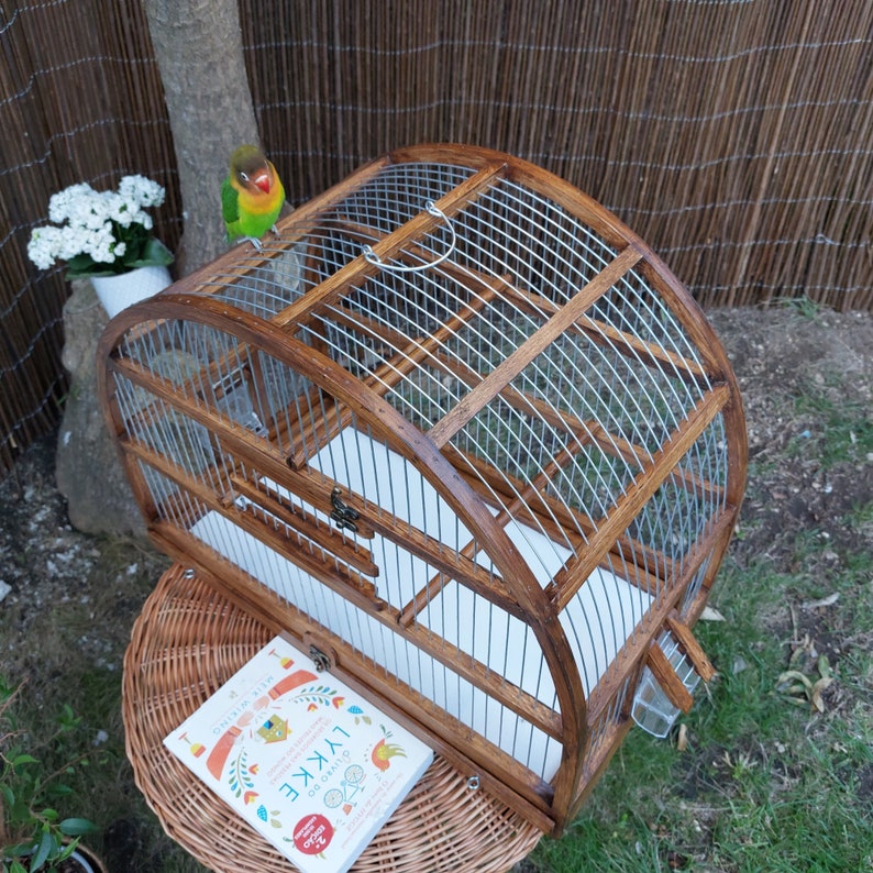May include: A wooden birdcage with a curved top and a metal wire grid. The cage is open and has a white paper lining. A small green and yellow bird is perched on the top of the cage.
