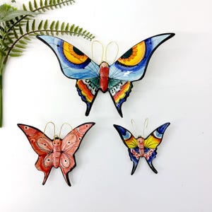Op de afbeelding: Drie keramische vlinder wanddecoraties. De grootste vlinder heeft blauwe vleugels met gele en oranje accenten. De andere twee vlinders zijn kleiner, één met roze en rode vleugels en de andere met blauwe en gele vleugels.