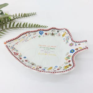 Op de afbeelding: Wit, bladvormig keramisch serveerbord met een rood-zwarte patroonrand. Het bord is versierd met kleurrijke bloemen- en hartmotieven en bevat handgeschreven tekst in meerdere kleuren.