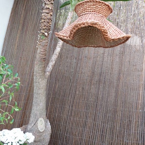 May include: A woven brown lampshade with a flared, ruffled edge hangs from a tree branch. The lampshade is made of natural fibers and has a woven pattern. The background features a brown bamboo fence and a tree trunk.