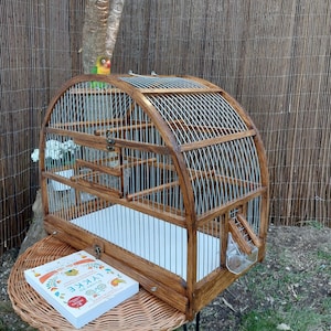 May include: A wooden birdcage with a curved top and a metal wire mesh. The cage has a white bottom and a small door on the side. The cage is sitting on a wicker table with a book on top. A green and yellow bird is perched on the top of the cage.