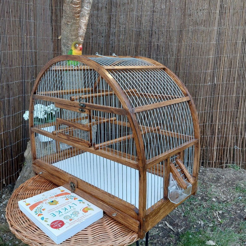 May include: A wooden birdcage with a curved top and a metal wire mesh. The cage has a white bottom and a small door on the side. The cage is sitting on a wicker table with a book on top. A green and yellow bird is perched on the top of the cage.