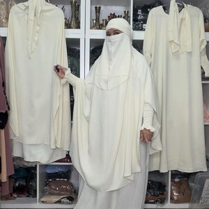 May include: An ivory-colored modest dress ensemble, including a long dress and a matching head covering. The outfit features flowing fabric and a modest design. The image shows the outfit on a person, with additional dresses hanging in the background.