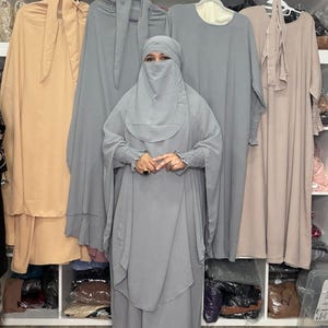 May include: A woman wearing a gray niqab and matching abaya, standing in front of a rack of clothing. Other garments in beige and gray hang on the rack. The abaya has long sleeves with elastic cuffs. The background includes shelves with decorative items.
