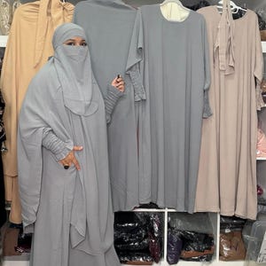 May include: A woman wearing a light gray abaya and niqab stands next to several hanging abayas in various colors. The abayas are displayed on white shelves. The sleeves of the abaya have a ruched design.