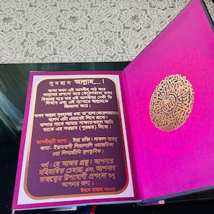 May include: An open book with a magenta cover and pages. The right page features gold calligraphy within a decorative circular design. The left page has text in a gold frame. A blue and red ribbon bookmark is visible.
