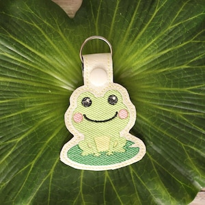 May include: A green frog with pink cheeks sitting on a green lily pad. The frog is embroidered on a white fabric keychain.