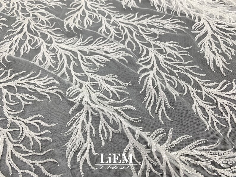 May include: White beaded lace fabric with a delicate floral pattern. The fabric is sheer and has a soft, flowing texture.