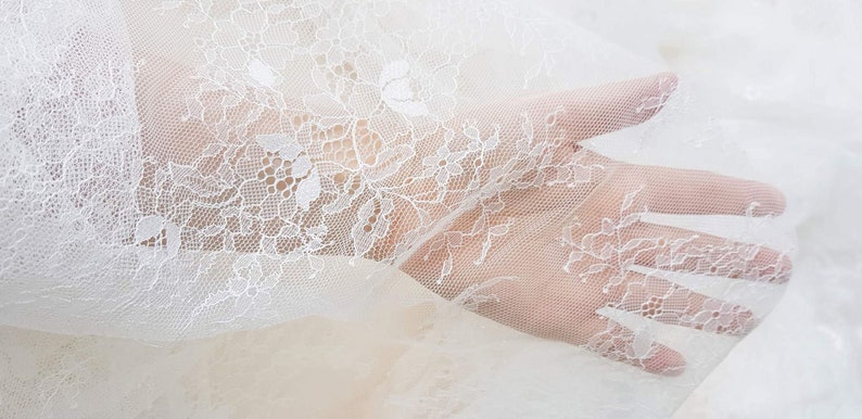 May include: White lace fabric with a delicate floral pattern. The fabric is sheer and has a soft, flowing texture.