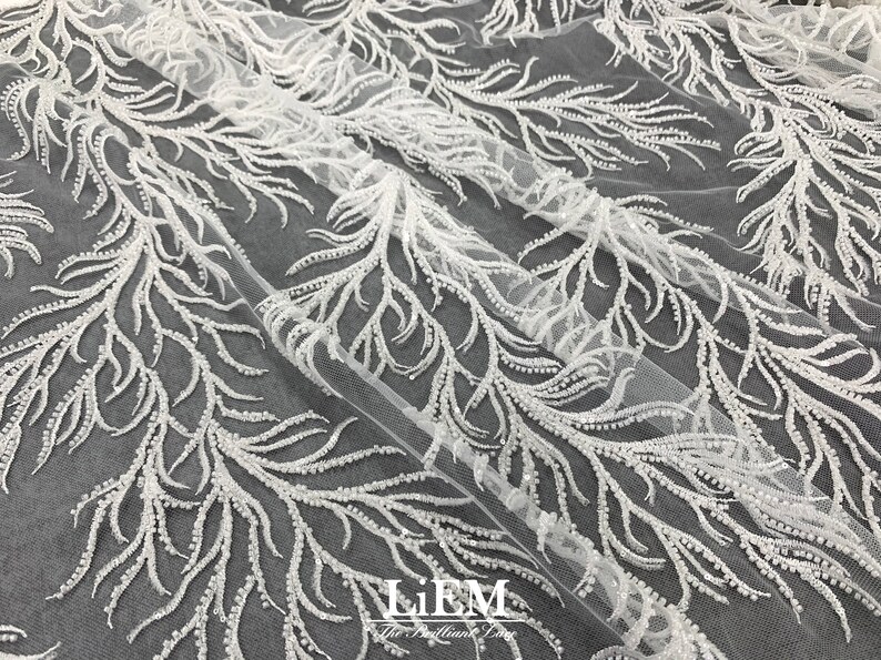 May include: White beaded lace fabric with a delicate feather design. The fabric is sheer and has a soft, flowing texture. The beads are small and evenly spaced, creating a subtle shimmer.