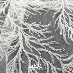 May include: White beaded lace fabric with a delicate feather design. The fabric is sheer and has a soft, flowing texture. The beads are small and evenly spaced, creating a subtle shimmer.