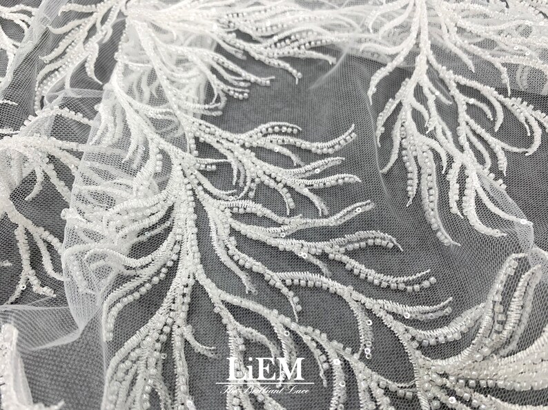 May include: White beaded lace fabric with a delicate feather design. The fabric is sheer and has a soft, flowing texture. The beads are small and evenly spaced, creating a subtle shimmer.