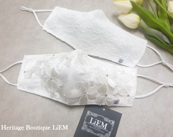 Puede incluir: Dos mascarillas blancas de encaje con delicado bordado floral y pedrería. Las mascarillas están atadas con una cinta de satén blanca. El texto "Heritage Boutique LiEM" es visible en la esquina inferior izquierda de la imagen. El texto "LiEM" es visible en la esquina inferior derecha de la imagen.