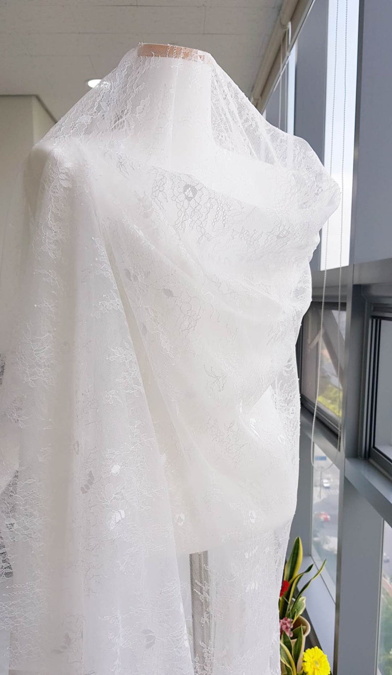May include: White lace fabric with a delicate floral pattern. The fabric is draped over a white mannequin.