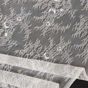 May include: White lace fabric with a delicate floral pattern. The lace is soft and sheer, and it has a subtle shimmer.
